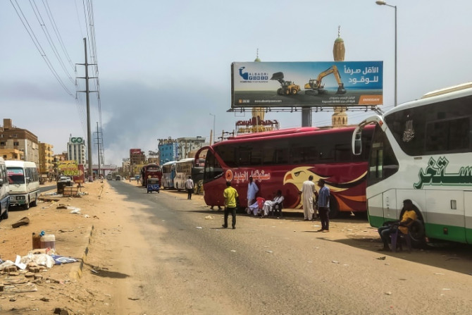 Fumaça à distância enquanto as pessoas esperam ao lado de ônibus de passageiros na capital do Sudão, Cartum